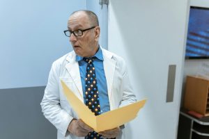 A senior doctor wearing eyeglasses examines a file in a hospital corridor.