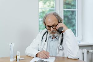 Senior doctor with gray hair writing notes during a telemedicine call.